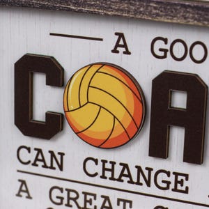 May include: A white wooden sign with dark brown raised lettering and a central graphic of a yellow and orange volleyball. The text reads A GOOD COACH CAN CHANGE A GAME A GREAT COACH CAN CHANGE A LIFE, with the volleyball replacing the letter O in COACH.