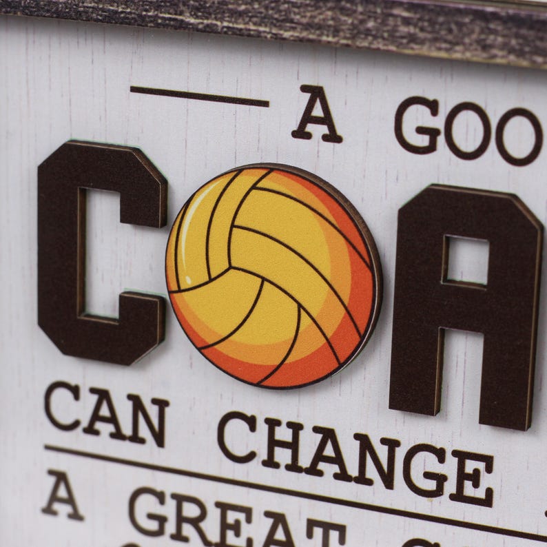 May include: A white wooden sign with dark brown raised lettering and a central graphic of a yellow and orange volleyball. The text reads A GOOD COACH CAN CHANGE A GAME A GREAT COACH CAN CHANGE A LIFE, with the volleyball replacing the letter O in COACH.