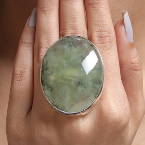 May include: A large oval ring featuring a polished, translucent pale green stone with internal cloudy inclusions and dark flecks, set within a smooth, shiny silver-toned metal bezel that encircles the entire perimeter of the gemstone.