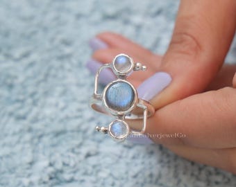 May include: A silver ring featuring three iridescent, round gemstones. The largest stone is centered, with two smaller stones above and below. The ring has a unique, geometric design with curved and straight lines. The gemstones have a blue and white color.