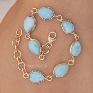 May include: A gold-toned chain bracelet featuring six teardrop-shaped light blue Larimar stones set in metal bezels. The bracelet includes a lobster claw clasp and a series of circular links for adjustment. The text ElegantSilverJewelCo is visible.