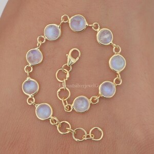 Könnte beinhalten: Ein goldfarbenes Armband mit runden, irisierenden Mondstein-Cabochons. Das Armband hat ein filigranes Kettendesign und einen Karabinerverschluss. Die Mondsteine sind in einzelnen Fassungen gefasst, was einen eleganten Look erzeugt. Das Armband ist ca. 18 cm lang.