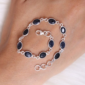 May include: A silver chain bracelet featuring nine oval-shaped, faceted dark blue sapphire gemstones set in individual metal bezels. The bracelet includes a lobster claw clasp and a short extender chain with circular links at the end for adjustment.