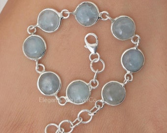 May include: A silver chain bracelet featuring seven round, pale blue cabochon stones set in smooth metal bezels. The bracelet includes a lobster claw clasp and an adjustable extender chain with circular links, creating a delicate and simple design.