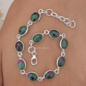 Puede incluir: Un brazalete de plata con un diseño de cadena de eslabones y piedras preciosas verdes de forma ovalada. El brazalete tiene un cierre de langosta.