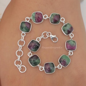 May include: A silver bracelet with a chain link clasp features square-shaped green and pink gemstones. The bracelet is on a person's wrist. "ElegantSilverJewelCo" is printed on the bracelet.