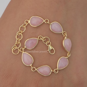 May include: A gold-toned chain bracelet featuring six smooth, teardrop-shaped light pink stones set in gold-toned bezels. The bracelet includes a lobster claw clasp and a short extender chain with larger circular links for adjustable sizing.