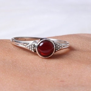 May include: A silver ring featuring a round, deep red garnet gemstone set in the center. The band has a split design on both sides of the stone, accented with small, raised metallic bead details that add texture to the polished silver surface.