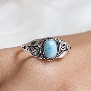 May include: A silver ring featuring a central oval-shaped light blue Larimar stone. The stone is set within a twisted rope-style border, flanked by decorative silver metalwork that includes swirling scroll patterns and leaf-like shapes on both sides.