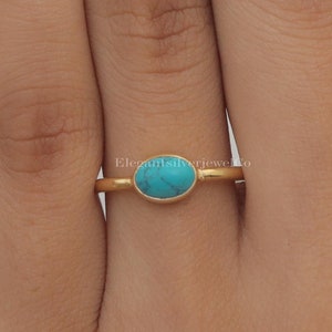Könnte beinhalten: Ein goldfarbener Ring mit einem kleinen, ovalen Türkisstein mit dunkler Maserung, der in einer glatten Fassung sitzt. Die Ringschiene ist dünn und poliert. Der Text "ElegantsilverjewelCo" ist in der Mitte des Rings und auf dem Finger sichtbar.