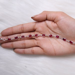May include: A silver chain bracelet featuring ten oval-shaped, deep red gemstones set in individual metal bezels. The bracelet includes a small lobster claw clasp at one end and a series of circular jump rings at the other for adjustable sizing.