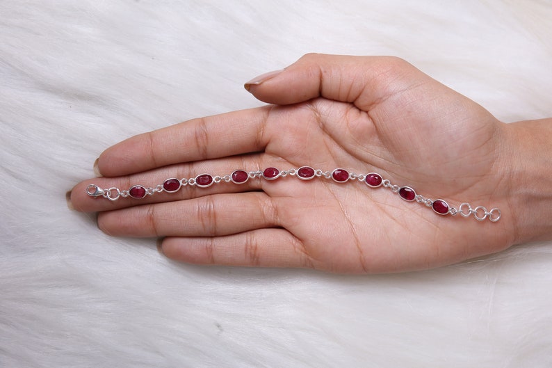 May include: A silver chain bracelet featuring ten oval-shaped, deep red gemstones set in individual metal bezels. The bracelet includes a small lobster claw clasp at one end and a series of circular jump rings at the other for adjustable sizing.