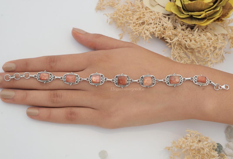 May include: A silver link bracelet featuring seven rectangular sunstone cabochons set in ornate metal frames. The bracelet includes a lobster claw clasp and a series of circular jump rings for adjustable sizing, showcasing a metallic, shimmering orange-brown stone texture.