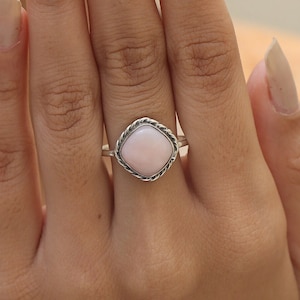 May include: A silver ring featuring a smooth, square-shaped light pink stone set in a twisted rope-style metal border. The band is thin and metallic, holding the central gemstone securely in place with a decorative, textured frame around the edges.