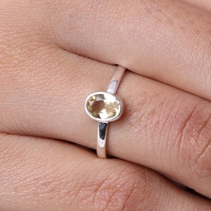 Peut inclure: Une bague en argent ornée d'une pierre précieuse ovale de couleur jaune pâle, sertie dans une monture en métal lisse. L'anneau est fin et poli, présentant un design épuré qui met en valeur la clarté et les facettes réfléchissantes de la pierre.