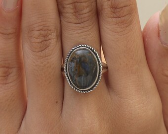 Puede incluir: Anillo ovalado con una banda plateada y una piedra texturizada de color azul oscuro y marrón. La piedra está engastada dentro de un borde retorcido en forma de cuerda. El anillo se lleva en un dedo.