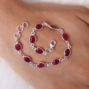 May include: A silver chain bracelet featuring multiple oval-shaped, faceted deep red ruby gemstones set in individual metal bezels. The bracelet includes a lobster claw clasp and a short extender chain with circular links at the end for adjustable sizing.