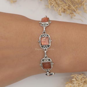 May include: A silver link bracelet featuring three square-cut, shimmering orange sunstone gemstones set in ornate, filigree-style metal frames. The bracelet displays a repeating pattern of these stone settings connected by small, polished silver circular links.
