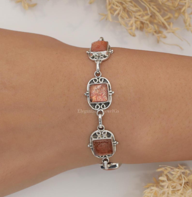 May include: A silver link bracelet featuring three square-cut, shimmering orange sunstone gemstones set in ornate, filigree-style metal frames. The bracelet displays a repeating pattern of these stone settings connected by small, polished silver circular links.