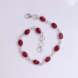 May include: A silver chain bracelet featuring nine oval-shaped, deep red gemstones set in individual silver bezels. The bracelet includes a lobster claw clasp and a short extender chain with circular links, creating a delicate and symmetrical design.