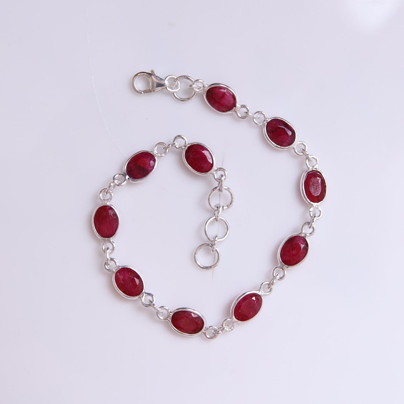 May include: A silver chain bracelet featuring nine oval-shaped, deep red gemstones set in individual silver bezels. The bracelet includes a lobster claw clasp and a short extender chain with circular links, creating a delicate and symmetrical design.