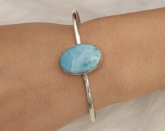 May include: A silver cuff bracelet featuring a large, polished, oval-shaped light blue Larimar gemstone set in a smooth metal bezel. The band of the bracelet is thin, polished, and metallic, creating a simple and clean design for the wrist.