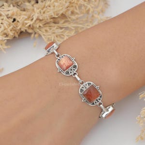 May include: A silver link bracelet featuring square-cut, shimmering orange sunstone gemstones. Each stone is set within an intricate, decorative silver frame with scrollwork details, connected by small silver links to form a continuous chain.