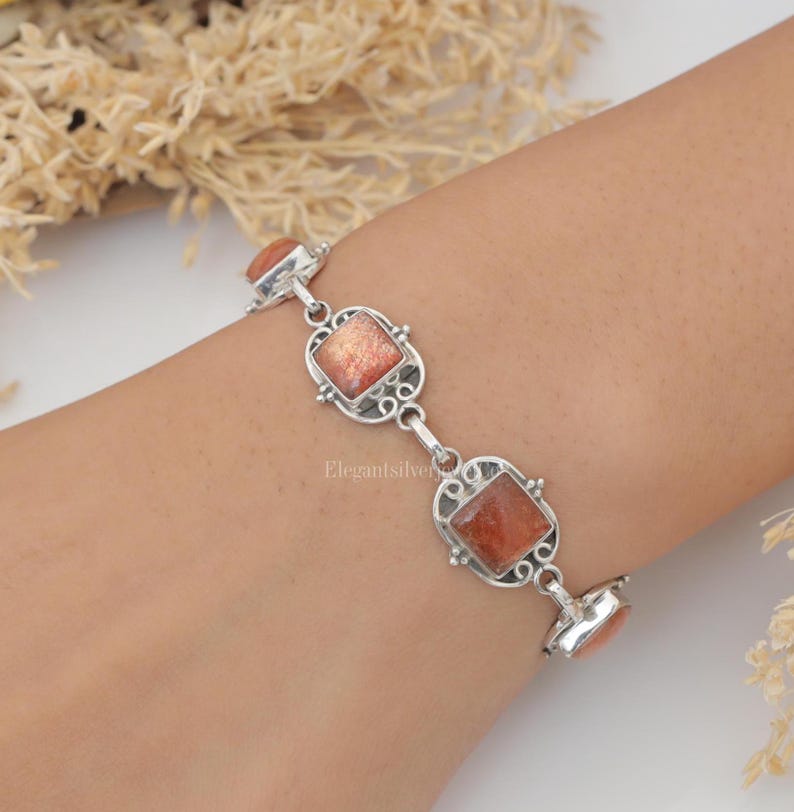 May include: A silver link bracelet featuring square-cut, shimmering orange sunstone gemstones. Each stone is set within an intricate, decorative silver frame with scrollwork details, connected by small silver links to form a continuous chain.