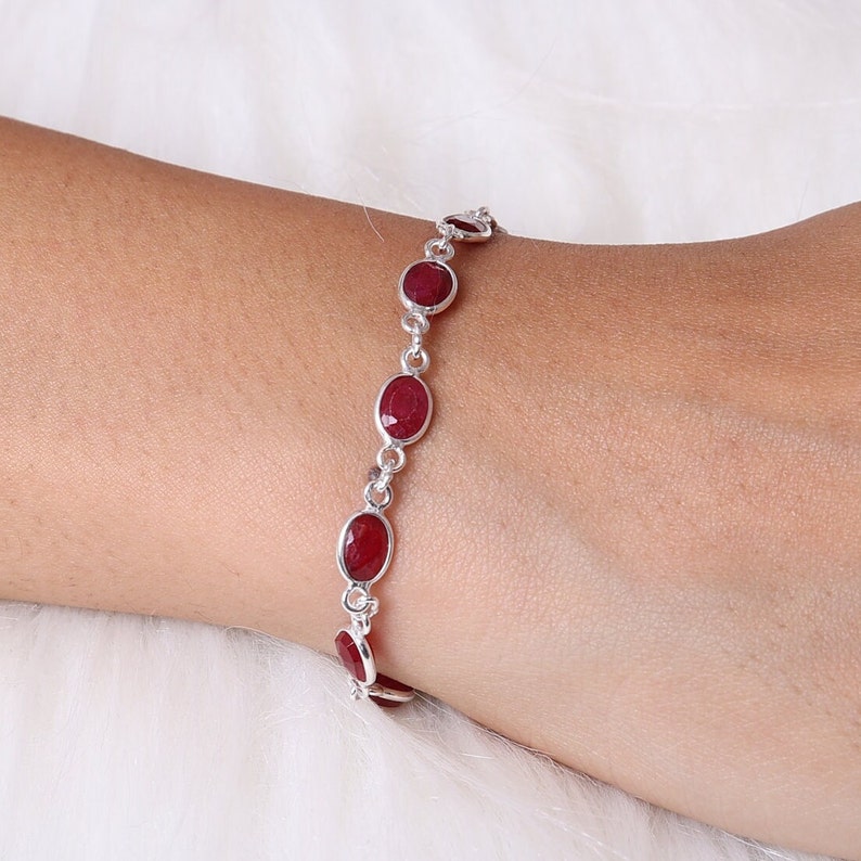 May include: A silver chain bracelet featuring a series of faceted, oval-shaped deep red gemstones set in polished metal bezels. The links connect each stone in a continuous line, creating a delicate and refined piece of jewelry for the wrist.