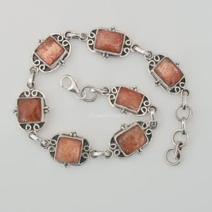 May include: A silver link bracelet featuring six rectangular, shimmering orange sunstone cabochons set in ornate, swirling metal frames. The bracelet includes a lobster claw clasp and a short extender chain with circular links for adjustable sizing.