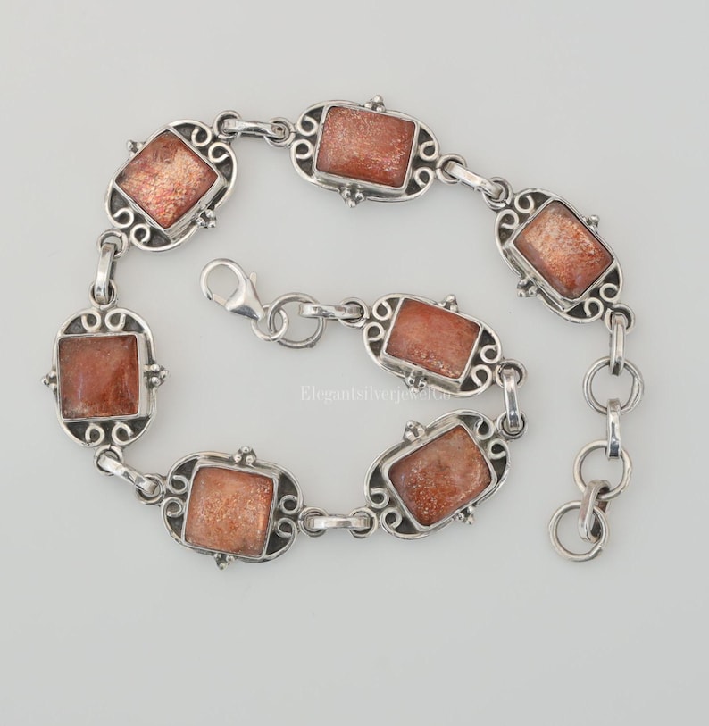 May include: A silver link bracelet featuring six rectangular, shimmering orange sunstone cabochons set in ornate, swirling metal frames. The bracelet includes a lobster claw clasp and a short extender chain with circular links for adjustable sizing.
