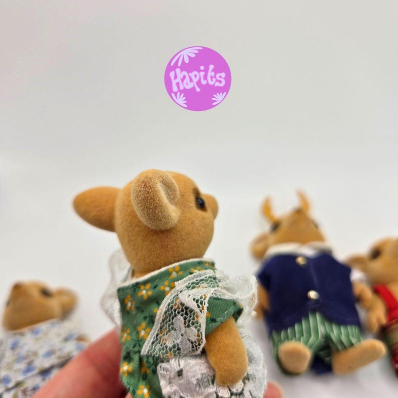 May include: A group of miniature animal figurines, including a bear-like figure in a green floral dress with white lace trim. Other figures are dressed in blue and red outfits. The image includes a purple circle with the text "Hapiks".