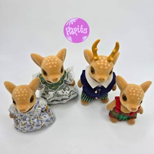 May include: Four fawn-colored deer figurines with white spotted patterns. Two deer wear dresses, one in blue floral and the other in green lace. One deer wears a navy blue vest and striped pants, and the last deer wears a red and green plaid outfit.