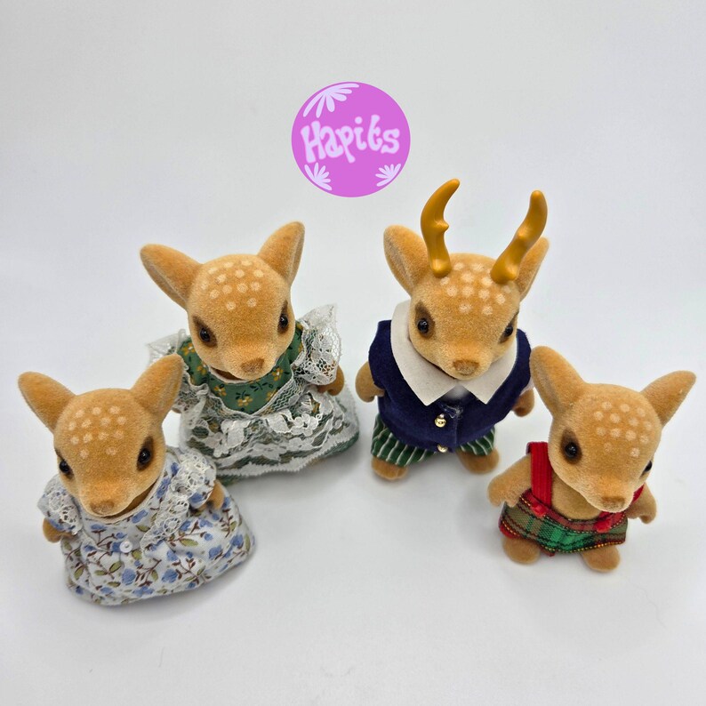 May include: Four fawn-colored deer figurines with white spotted patterns. Two deer wear dresses, one in blue floral and the other in green lace. One deer wears a navy blue vest and striped pants, and the last deer wears a red and green plaid outfit.