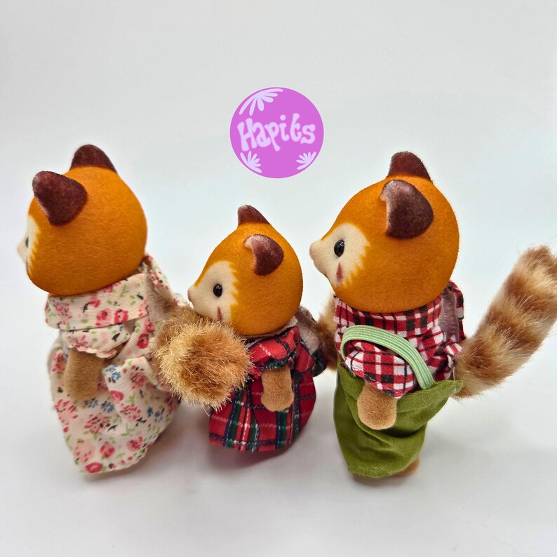 Puede incluir: Tres figuras de tanuki con cabezas naranjas y orejas marrones. Visten diferentes atuendos: vestido floral, camisa a cuadros rojos y pantalones verdes. Las figuras tienen un dise&ntilde;o lindo y de dibujos animados, con un logo morado en el fondo.