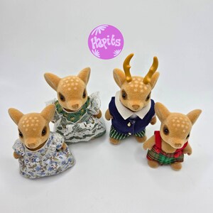 May include: Four fawn-colored deer figurines with white spots. Two deer wear floral dresses, one wears a navy blue jacket with green striped pants, and one wears a red and green plaid outfit. The image includes a purple circle with the text "Hapits".