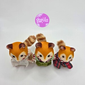 Puede incluir: Tres figuras de zorro de peluche con pelaje naranja, orejas marrones y colas a rayas. Cada zorro viste un atuendo diferente: floral, verde y a cuadros. La imagen incluye un c&iacute;rculo morado con el texto "Hapiks".