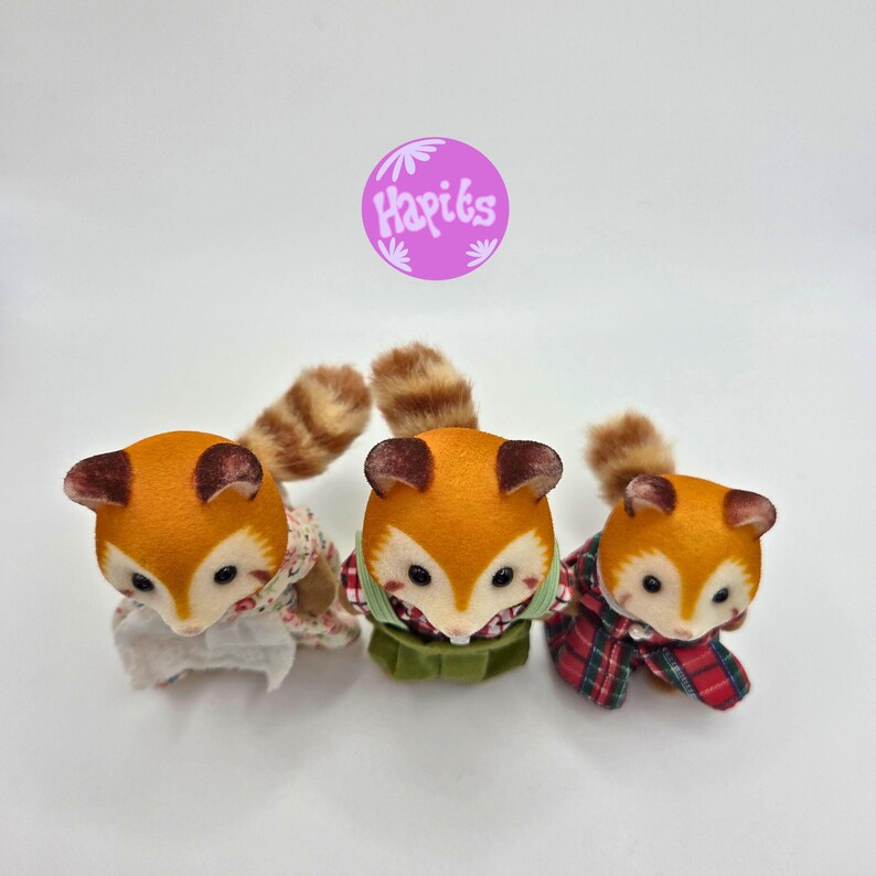 Puede incluir: Tres figuras de zorro de peluche con pelaje naranja, orejas marrones y colas a rayas. Cada zorro viste un atuendo diferente: floral, verde y a cuadros. La imagen incluye un c&iacute;rculo morado con el texto "Hapiks".