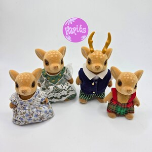 May include: Four fawn-colored deer figurines, each with a unique outfit. One wears a floral dress, another a green dress with lace, a third a navy vest and striped pants, and the last a plaid overall. A purple circle with the text "Hapits" is in the background.