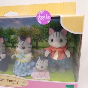 May include: A family of four gray and black tabby cats in a box. The father cat is wearing a red shirt with a yellow tie, the mother cat is wearing a yellow shirt with a blue bow and a blue and white skirt, the son cat is wearing a blue shirt and blue pants, and the daughter cat is wearing a pink dress with lace.