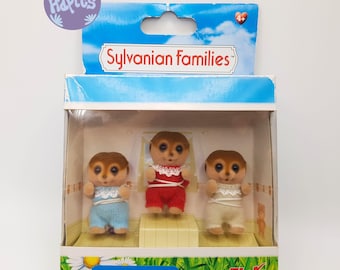 May include: A boxed set of Sylvanian Families Meerkat Triplets. The meerkats wear blue, red, and cream-colored outfits with white lace trim. The box features a sky and cloud background with the text "Sylvanian Families" and "Meerkat Triplets".