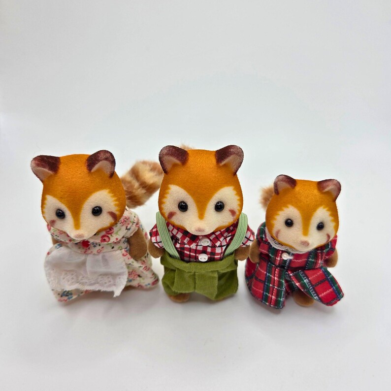 Puede incluir: Tres figuras de zorro con pelaje naranja, caras blancas y ojos oscuros. Los zorros visten trajes en miniatura: un vestido floral, una camisa a cuadros rojos con peto verde y un abrigo a cuadros rojos. Una encantadora colecci&oacute;n de criaturas del bosque.