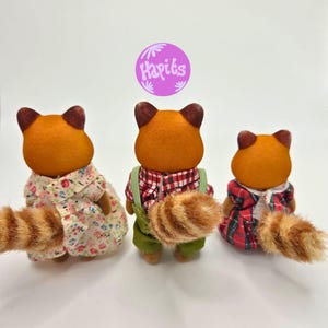 Puede incluir: Tres figuras de tanuki con pelaje naranja y orejas marrones. Visten diferentes atuendos: un vestido floral, una camisa a cuadros roja con peto verde y un vestido a cuadros rojo. Cada uno tiene una cola rayada. La imagen incluye un c&iacute;rculo morado con el texto "Hapiks".