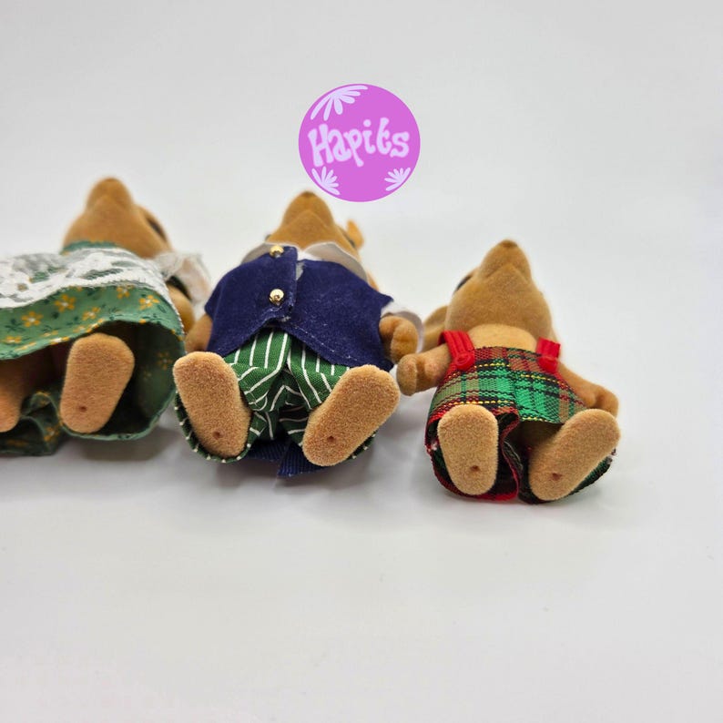 May include: Three miniature animal figurines, each dressed in different outfits. One wears a green and white dress, another a blue vest and striped pants, and the last a red plaid overall. The image includes the text "Hapiks" in a purple circle.
