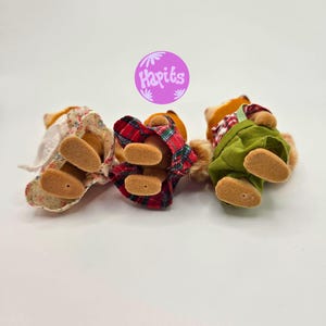Puede incluir: Tres peque&ntilde;as figuras de osos de peluche tumbadas boca arriba. Una lleva un vestido floral, otra un atuendo de cuadros rojos y la tercera un mono verde. Los osos tienen pelaje marr&oacute;n y pies color canela. Un c&iacute;rculo morado con el texto "Hapiks" est&aacute; en el fondo.