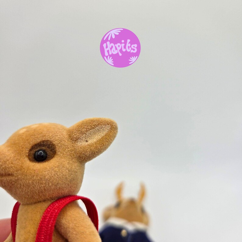 May include: A close-up of a tan deer figurine with a red strap. The deer has dark eyes and a detailed ear. In the background, a blurred figurine is wearing a blue jacket. A purple circle with the text "Hapiks" is in the upper right corner.