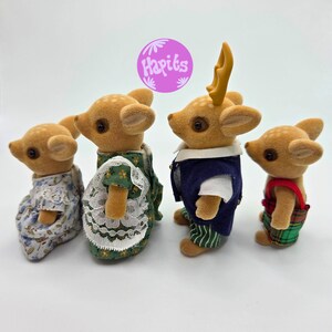 May include: Four miniature deer figurines, each with a unique outfit. The deer have brown fur and are wearing dresses, vests, and overalls. One deer has yellow antlers. The image includes a purple circle with the text "Hapiks".