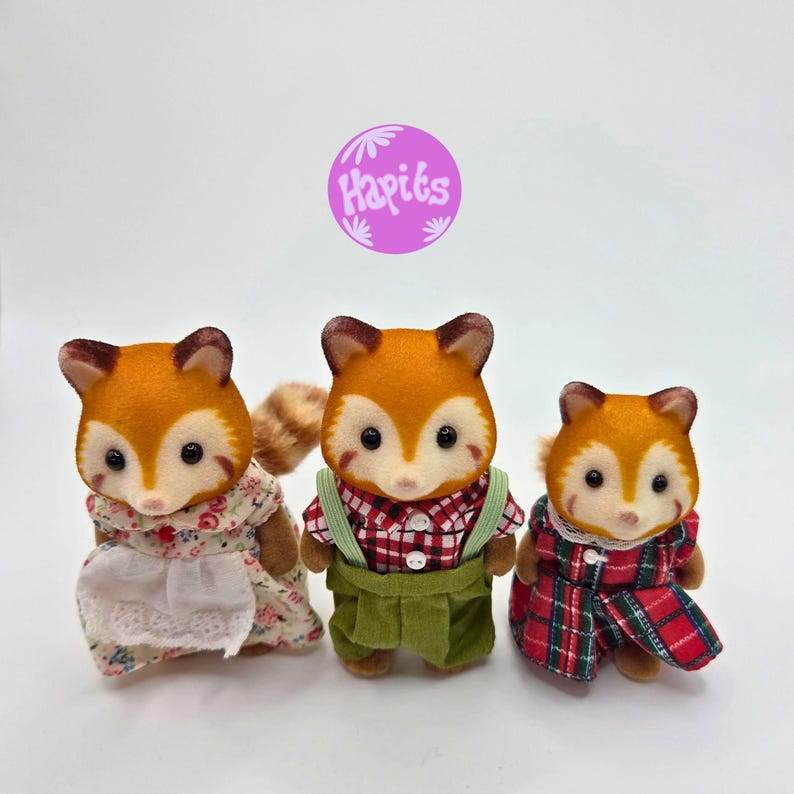 Puede incluir: Tres figuras de zorro con pelaje naranja y caras blancas. Los zorros visten diferentes atuendos: un vestido floral con delantal, una camisa a cuadros roja con peto verde y un vestido a cuadros rojo. La imagen incluye un logotipo morado con el texto "Hapiks".