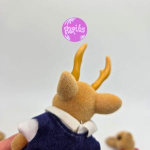 May include: A close-up of a small, tan-colored deer figurine with yellow antlers. The deer is wearing a navy blue vest with a white collar. The image includes a purple circle with the text "Hapiks".