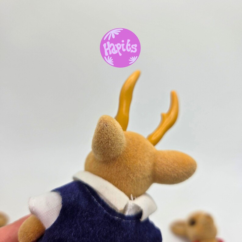May include: A close-up of a small, tan-colored deer figurine with yellow antlers. The deer is wearing a navy blue vest with a white collar. The image includes a purple circle with the text "Hapiks".
